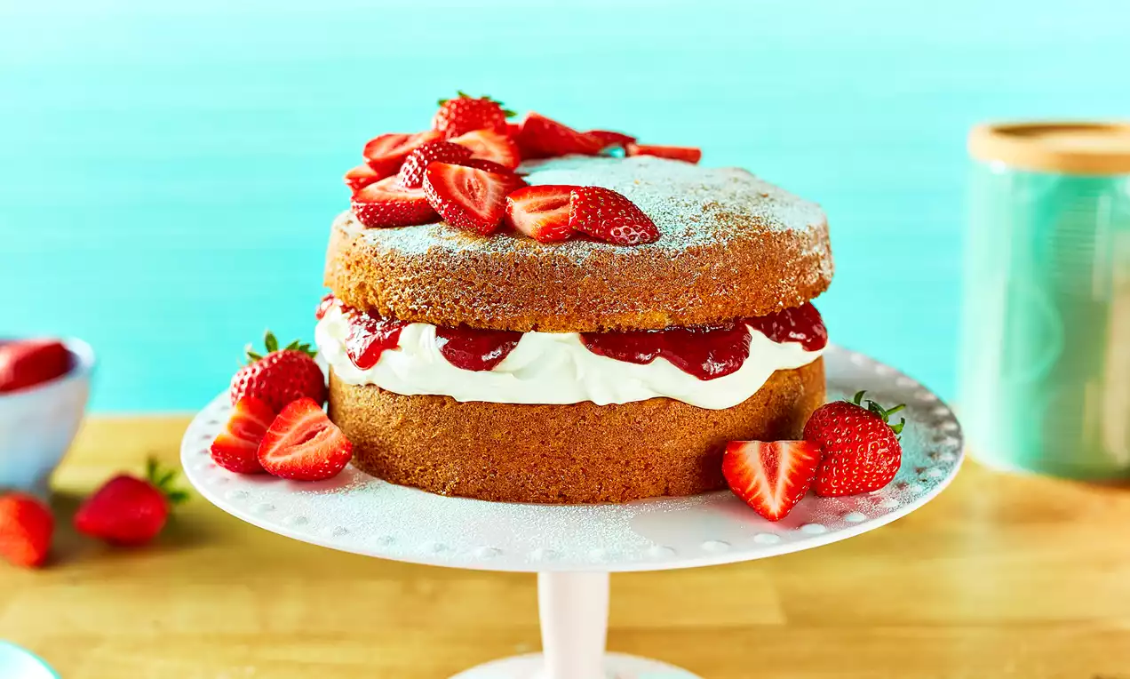Victoria sponge cake with golden sponge layers filled with whipped cream and red jam, topped with strawberries and a dusting of icing sugar