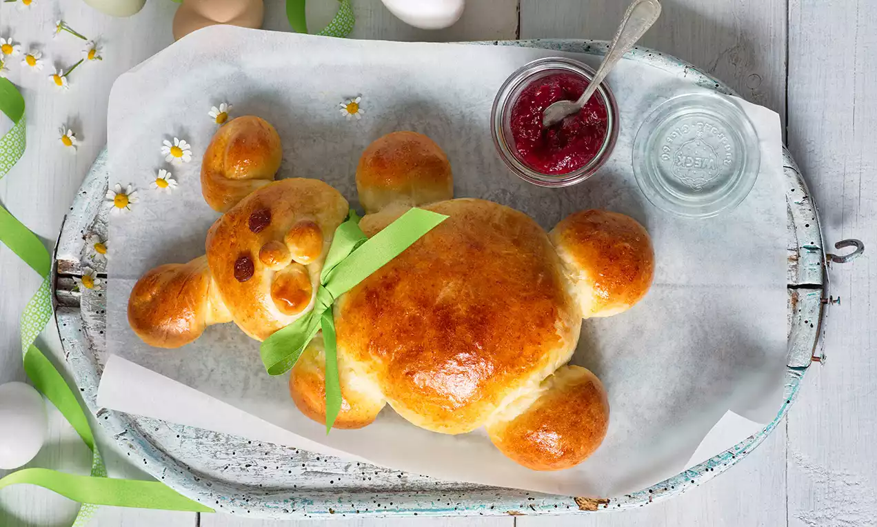 Zlatistý veľkonočný zajačik z kysnutého cesta s lesklým povrchom, zdobený zelenou stuhou, na plechu s papierom a miskami džemu a vody