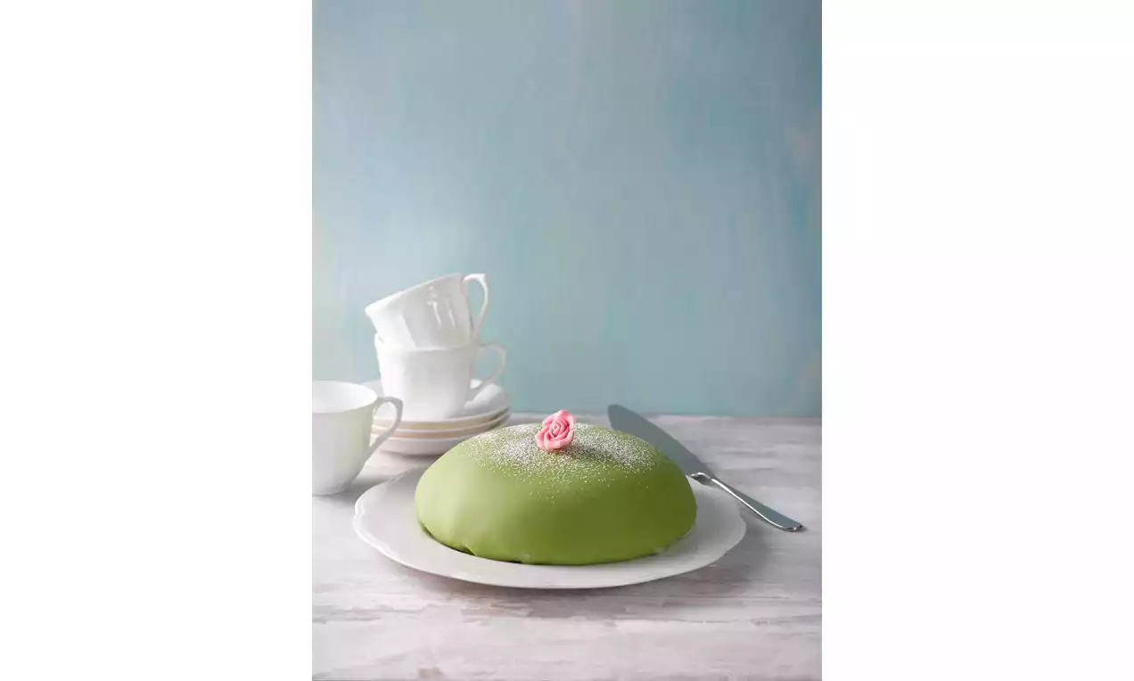 Vaaleanvihreällä marsipaanikuorella päällystetty kupolimainen kakku, jonka päällä sokeria ja vaaleanpunainen ruusukoriste valkoisella lautasella