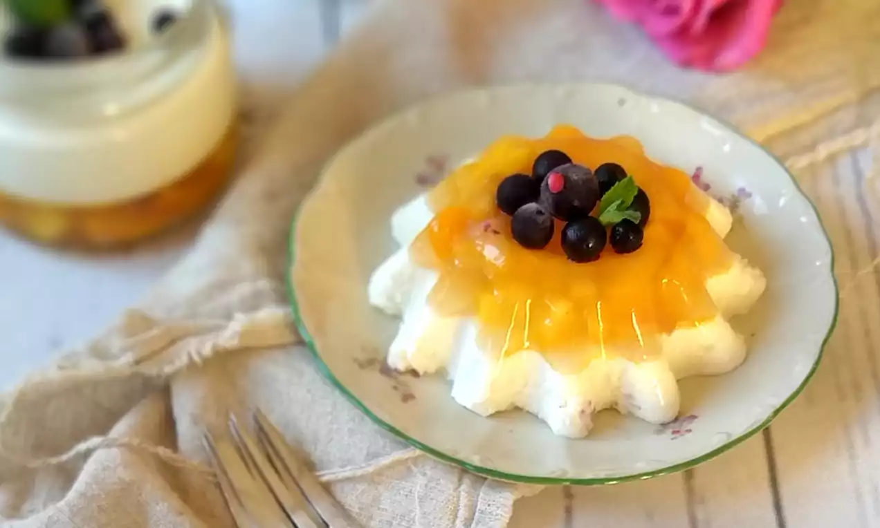 Panna cotta zamatovo bielej farby v tvare vlnkovanej kvetiny, pokrytá priezračným mangovým želé s čučoriedkami a lístkom mäty na kvietkovanom tanieri