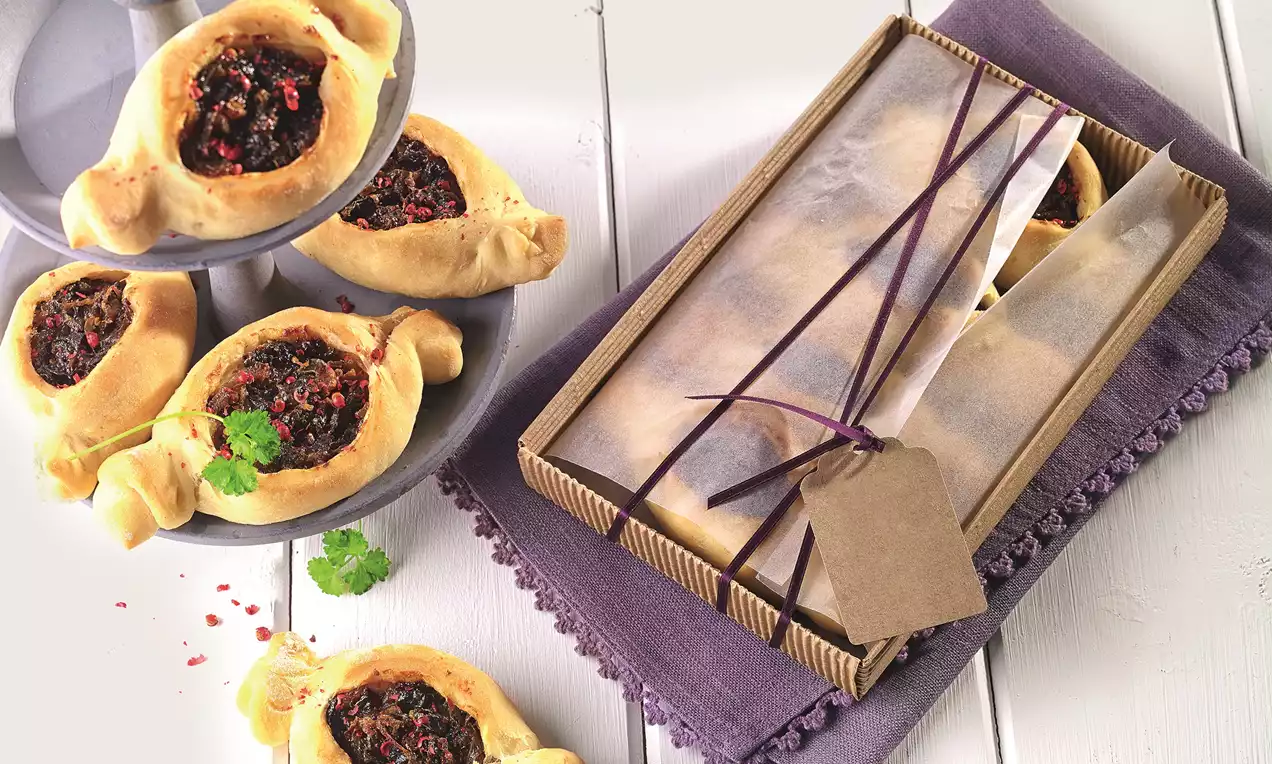 Zlatisté taštičky podlhovastého tvaru s tmavou slivkovou plnkou, ozdobené bylinkami a kúskami papriky, uložené na fialovom obruse a v krabičke