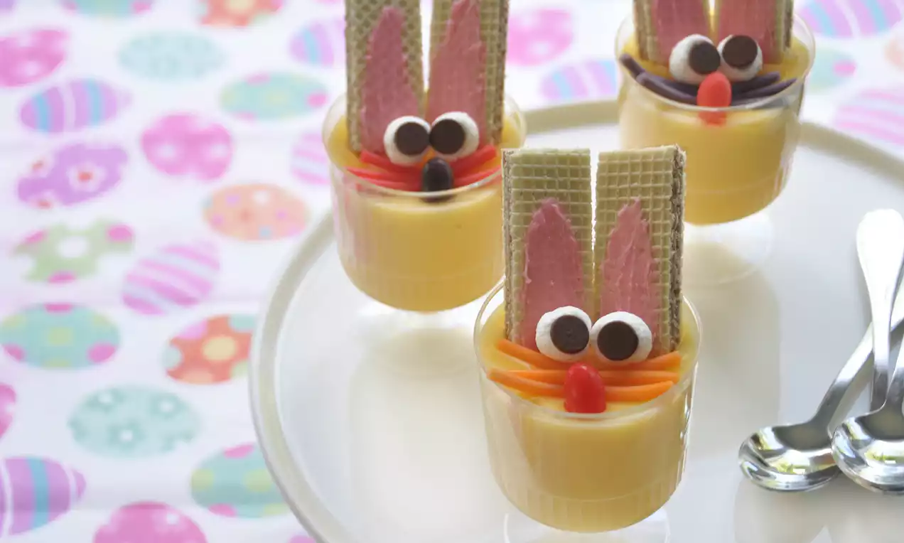 Small glass cups filled with pale yellow pudding, topped with wafer bunny ears, candy eyes, and colorful details on a white plate