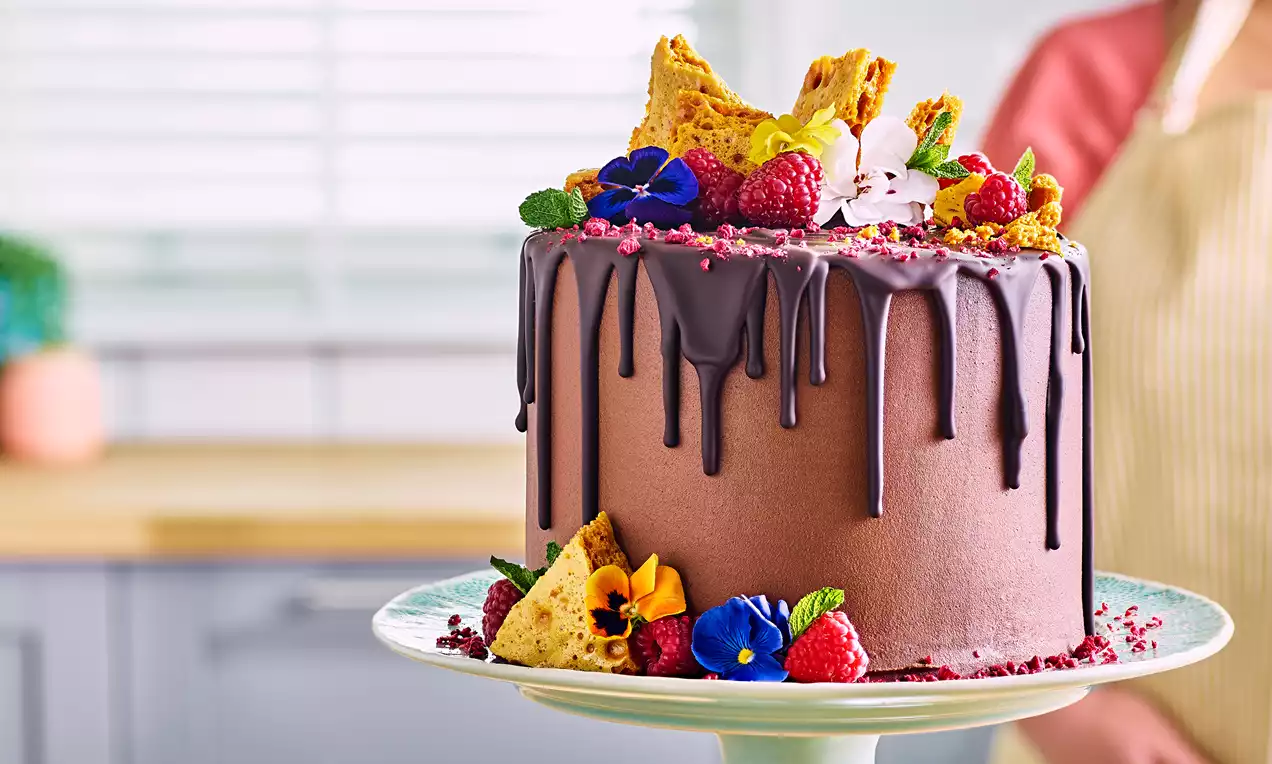 A round chocolate cake with glossy dark ganache drips, topped with honeycomb, raspberries, edible flowers, and set on a patterned plate