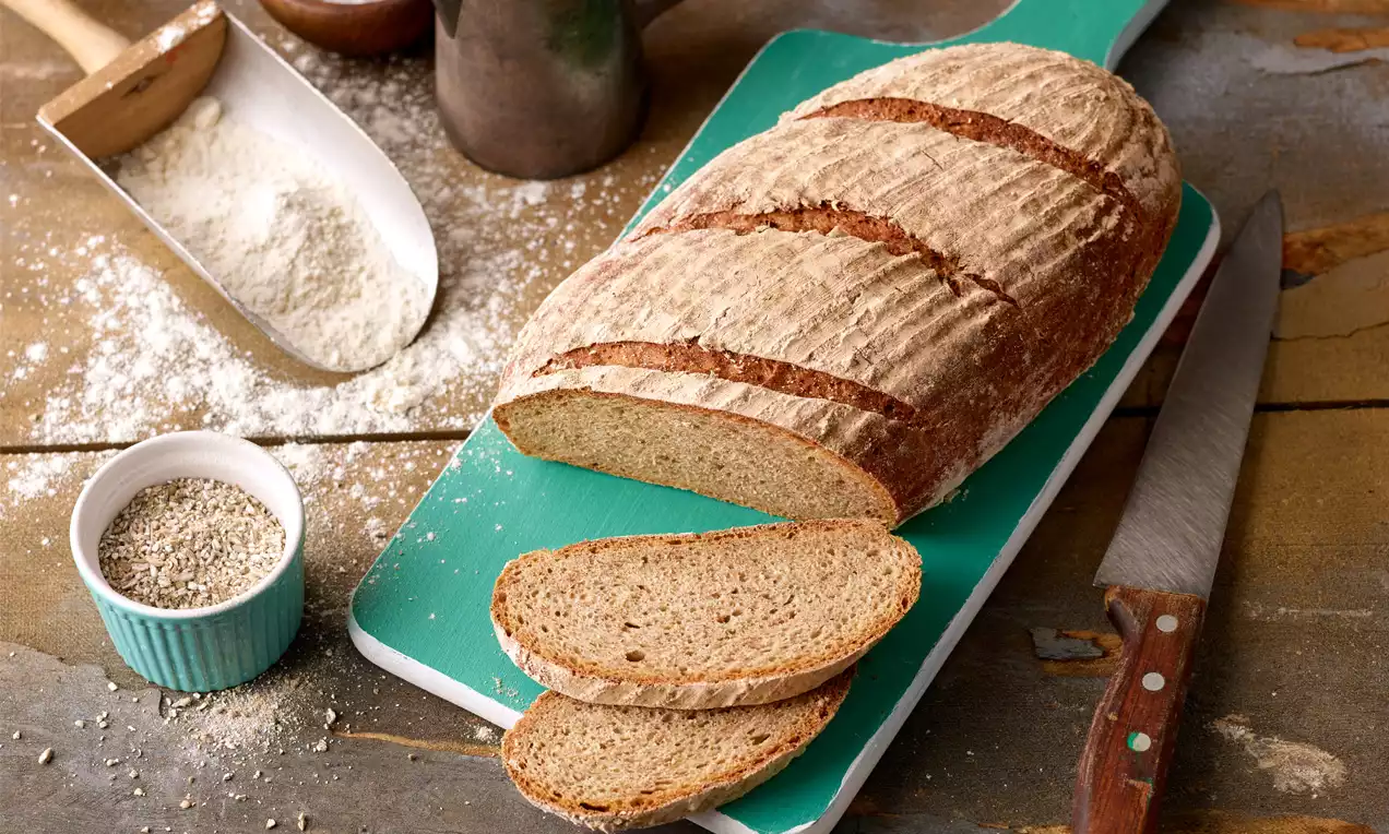 Roggenmischbrot mit bemehlter Kruste, länglich geformt, teils aufgeschnitten, auf grünem Brett mit Messer und Mehl auf Holztisch drapiert