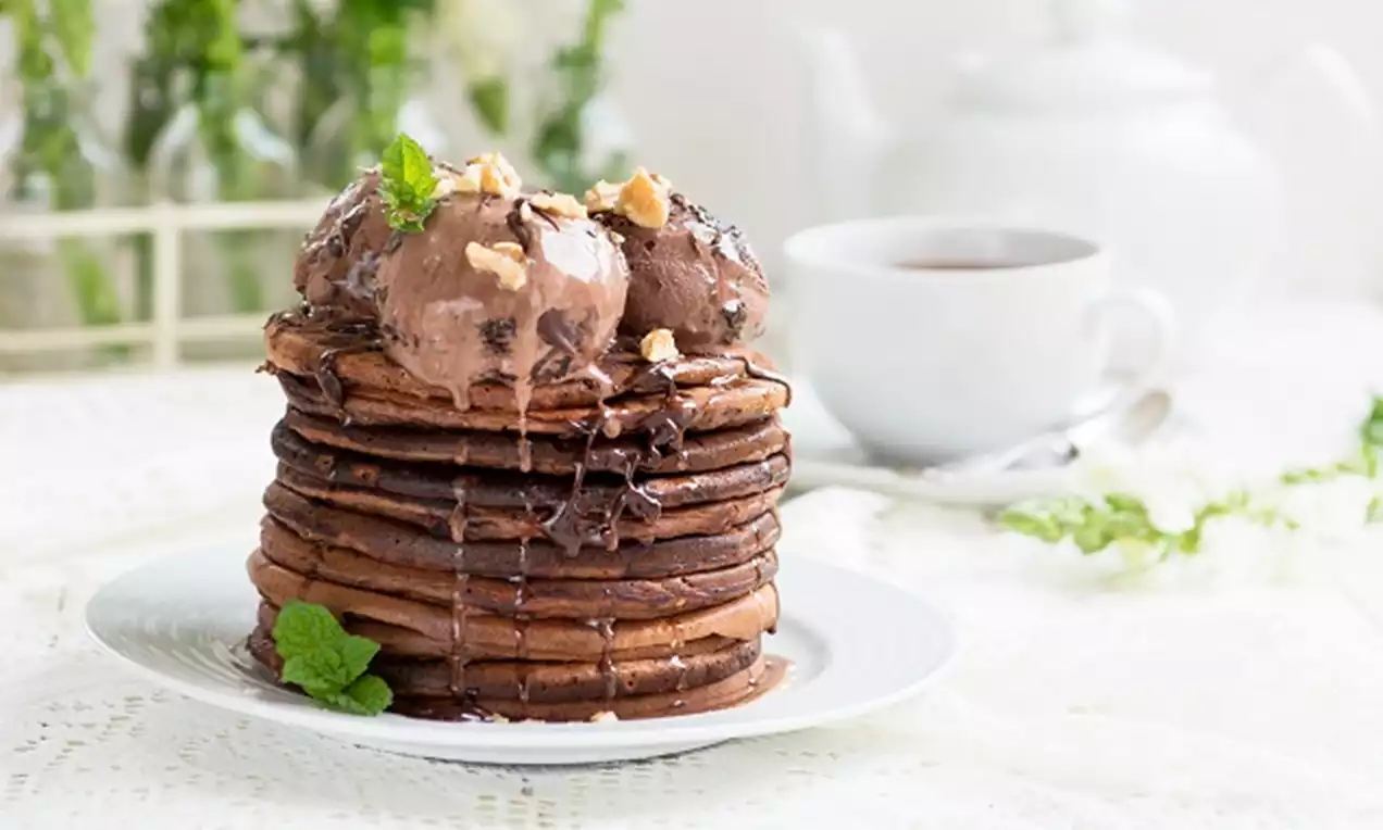 Stack of chocolate pancakes topped with scoops of chocolate ice cream, drizzled with chocolate sauce, garnished with mint and nuts on a white plate