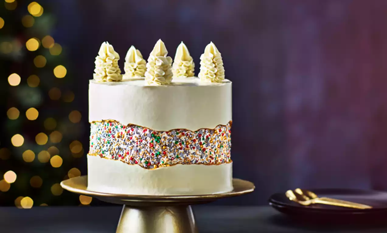 Tall white Christmas cake with a central band of vibrant rainbow sprinkles, topped with piped cream swirls, on a gold cake stand