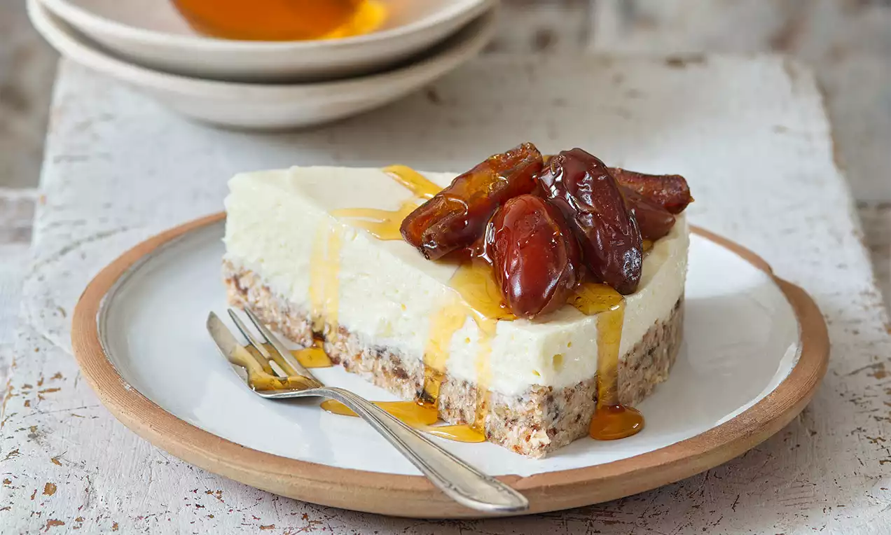 Šikmo nakrojený světlý dort s krémovou mascarpone vrstvou na hrubé oříškové základně, zdobený měkkými datlemi a zlatavým medem na bílém talíři