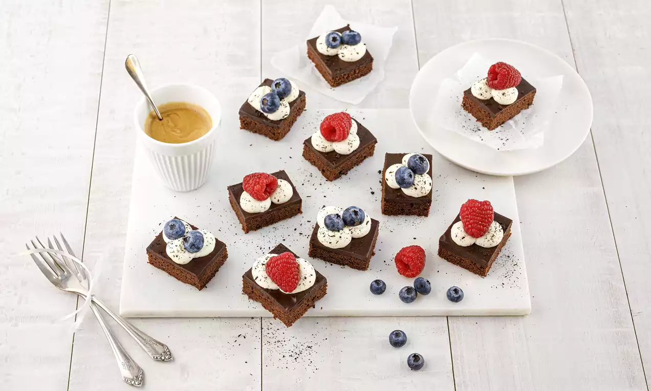 Quadratische Schokoschnitten mit weißer Sahnetupfen-Deko, Himbeeren und Blaubeeren auf hellem Untergrund und Beilagen angerichtet
