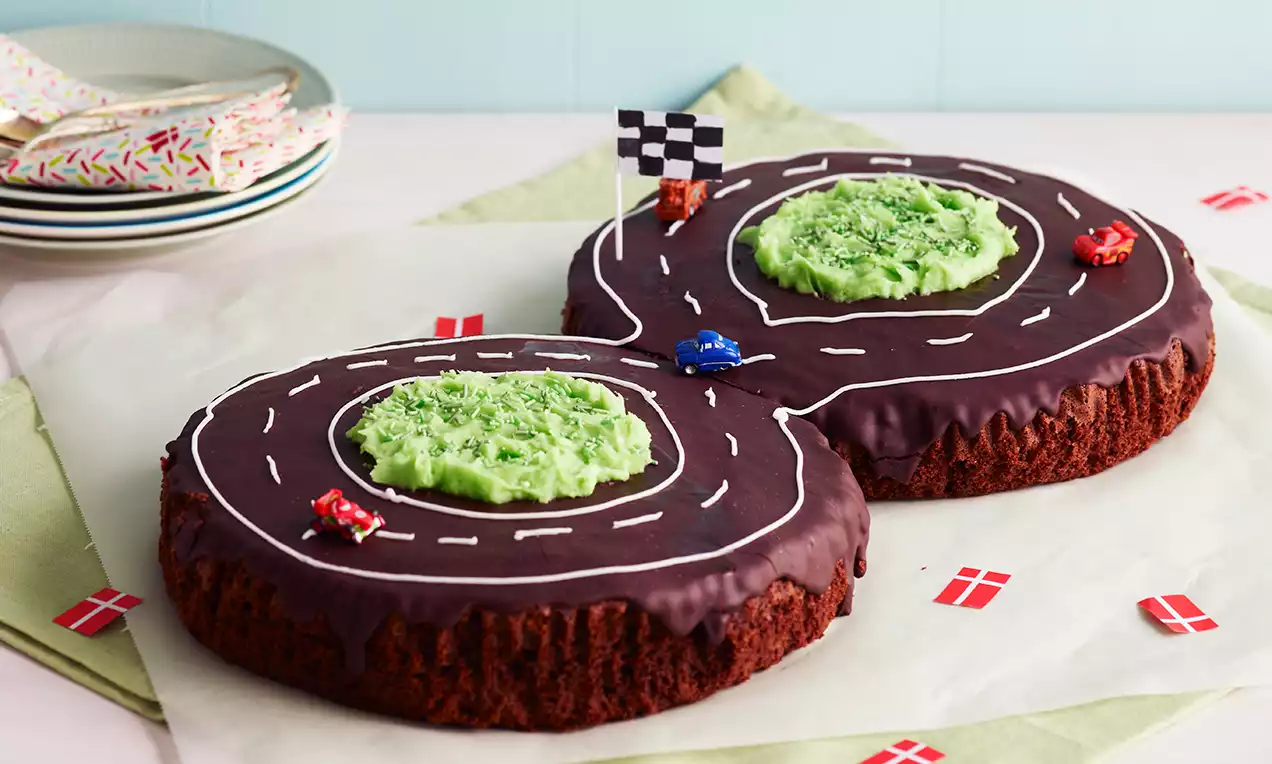Chokoladekage formet som en bilbane med mørk glasur, hvide striber og grønne græsområder, pyntet med små biler og flag