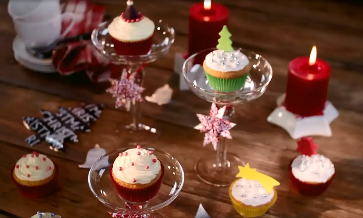Kerstcupcakes met romige witte topping, kleurrijke papieren vormpjes en decoraties, gepresenteerd op glazen standaarden met rode kaarsen