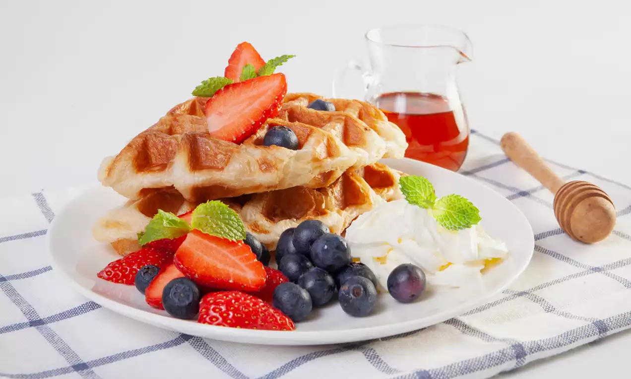 Golden, grid-patterned waffles stacked on a white plate with strawberries, blueberries, mint, whipped cream, and a jug of syrup beside