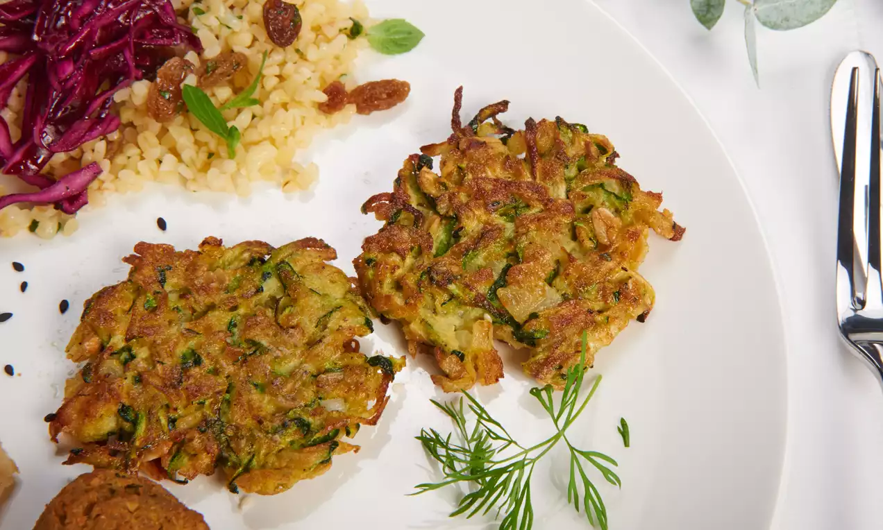 Goldbraune Zucchinipuffer mit feinen grünen Streifen und knuspriger Oberfläche, angerichtet auf einem weißen Teller mit frischen Kräutern
