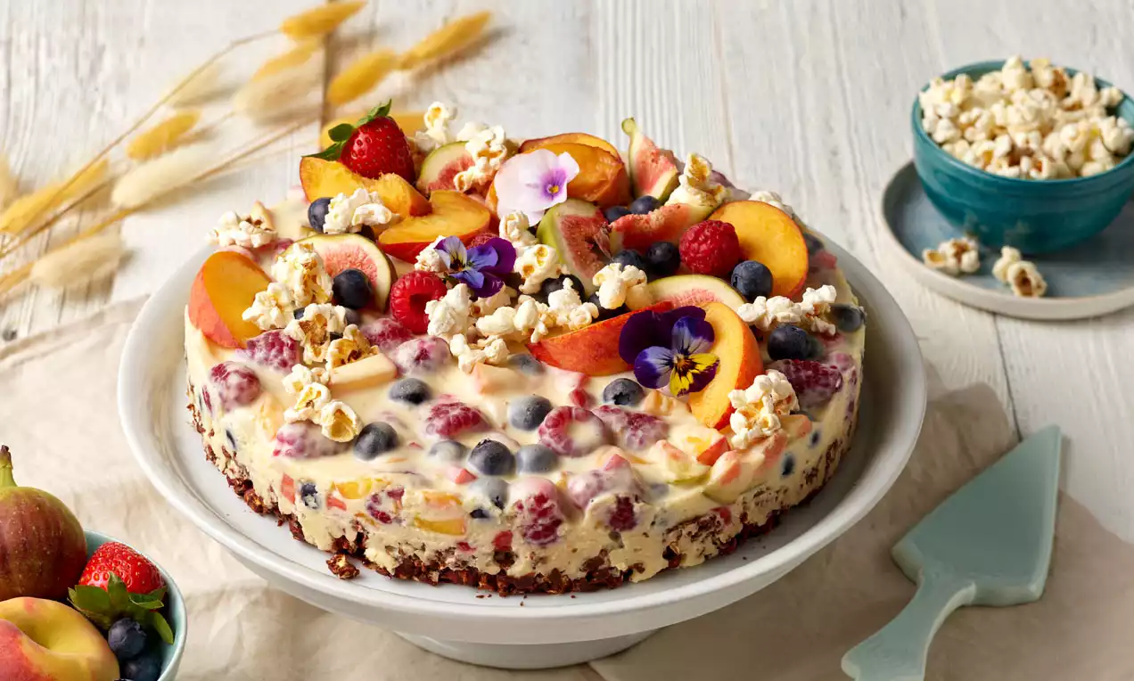 Bunte Torte mit saftigen Beeren, Pfirsichspalten, Popcorn und essbaren Blüten auf Nussboden auf weißer Tortenplatte angerichtet