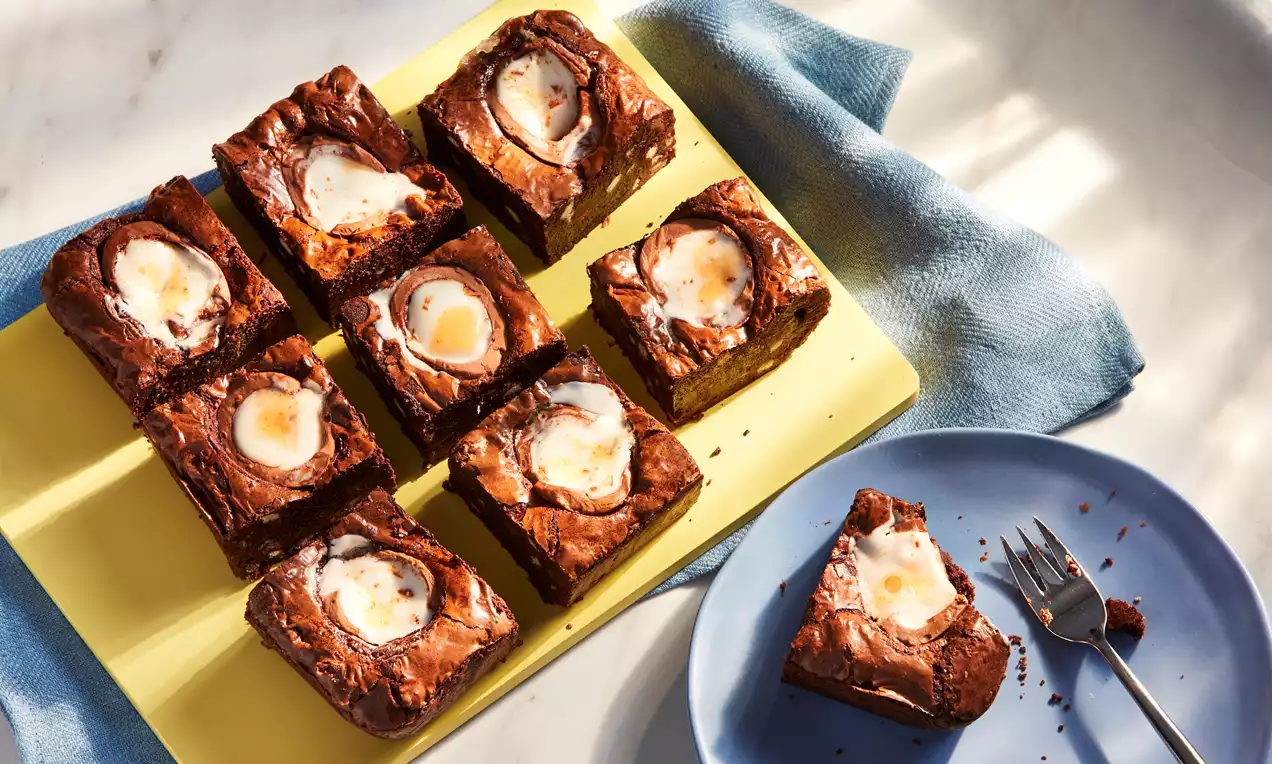 Rich chocolate brownies with a glossy top, each square featuring a halved Creme Egg, arranged on a yellow board near a blue plate