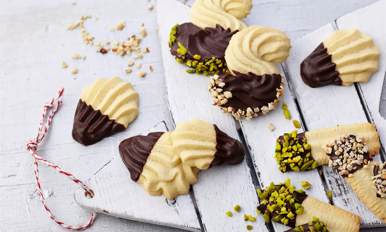 Helles, gerilltes Spritzgebäck, teils in dunkle Kuvertüre getaucht, mit gehackten Pistazien und Haselnüssen auf weißem Holzbrett arrangiert