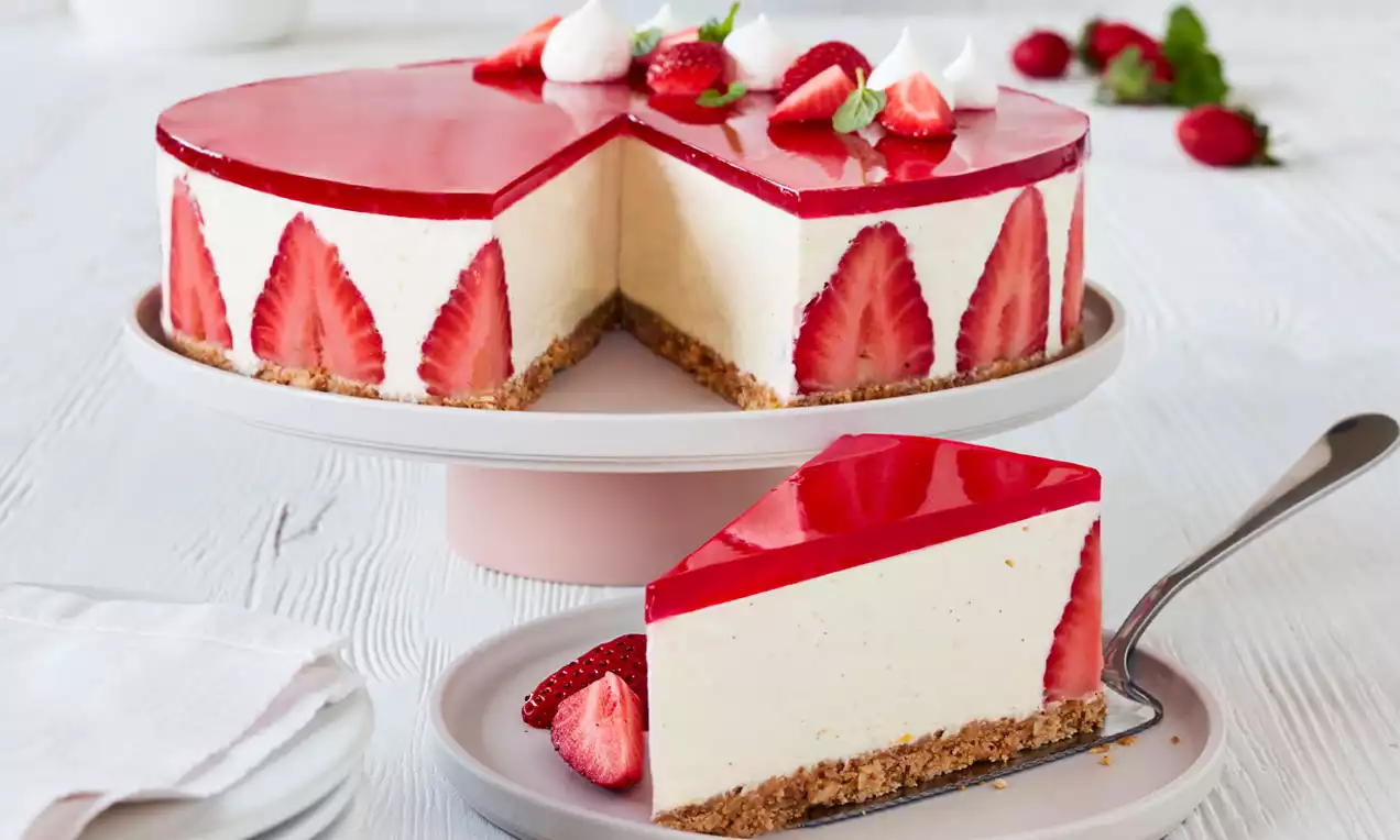 Cheesecake med lysebrunt kiksebund og cremefarvet fyld dekoreret med røde jordbærskiver og spejlblank jordbærgelé på hvidt kagefad