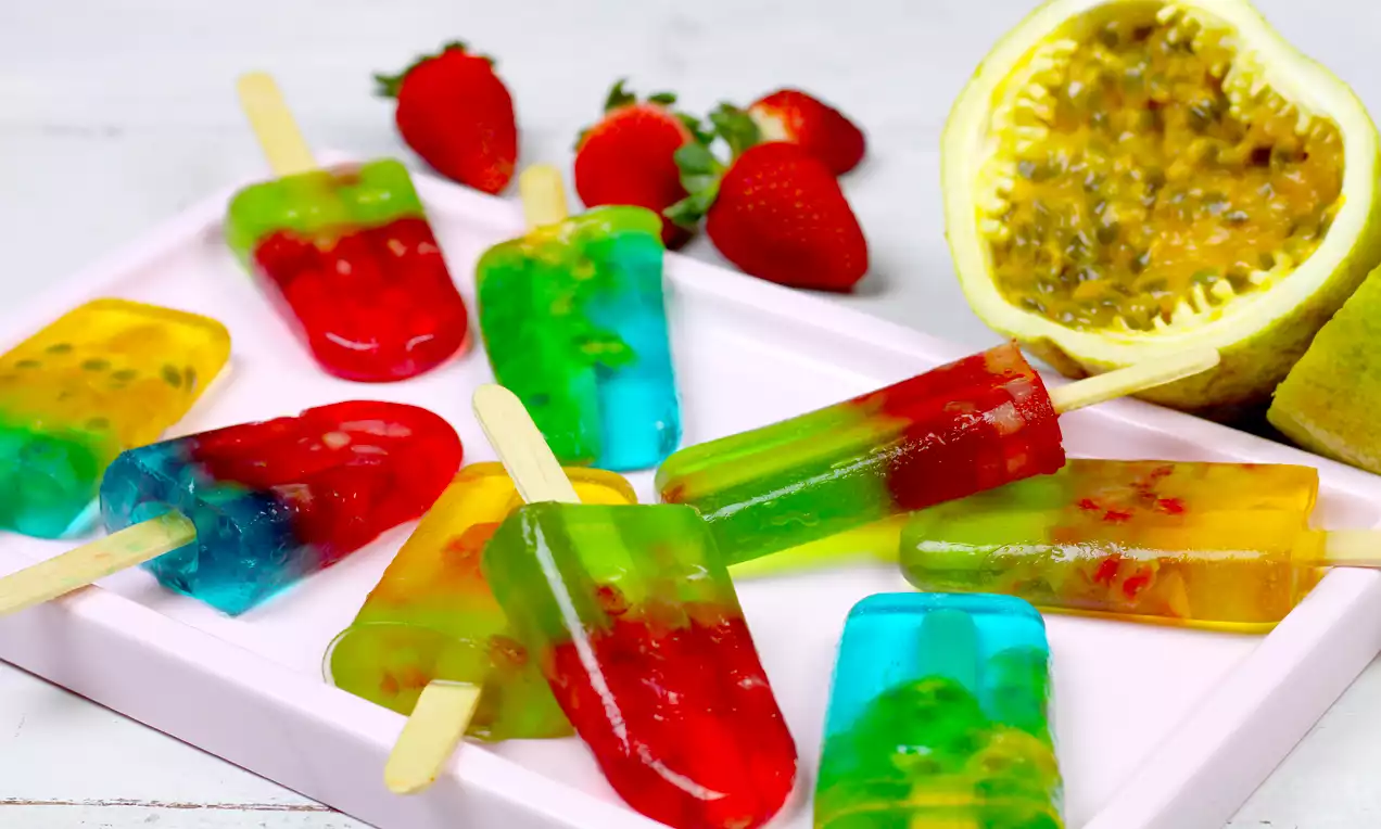 Picolés translúcidos de gelatina colorida com pedaços de frutas, em tons vibrantes de vermelho, verde, azul e amarelo sobre bandeja rosa