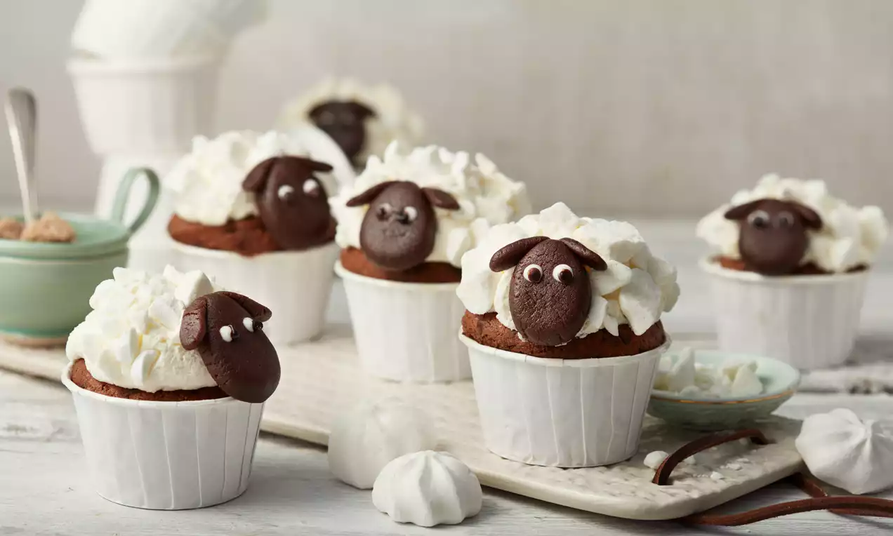 Čokoladni cupcakes u bijelim papirnatim košaricama, ukrašeni oblikovanim licima ovaca od tamne čokolade i bijelim hrpicama šlaga