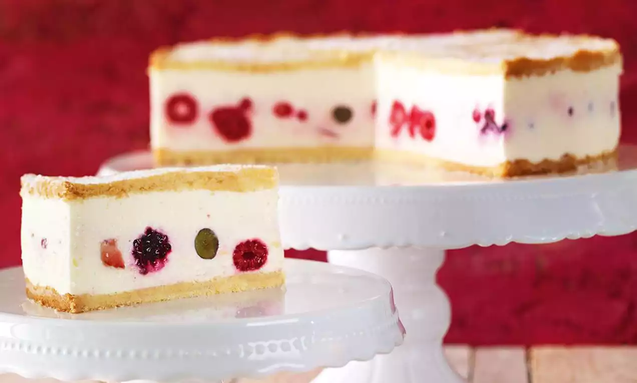 Tanka zlatnosmeđa korica zatvara slojeve svijetle kremaste mase prošarane cijelim bobičastim voćem, servirano na bijelom stalku