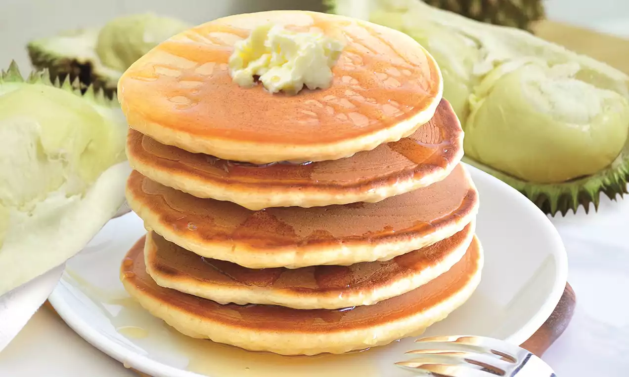 Golden pancakes stacked high, topped with a dollop of cream, drizzled with syrup on a white plate near peeled durian layers