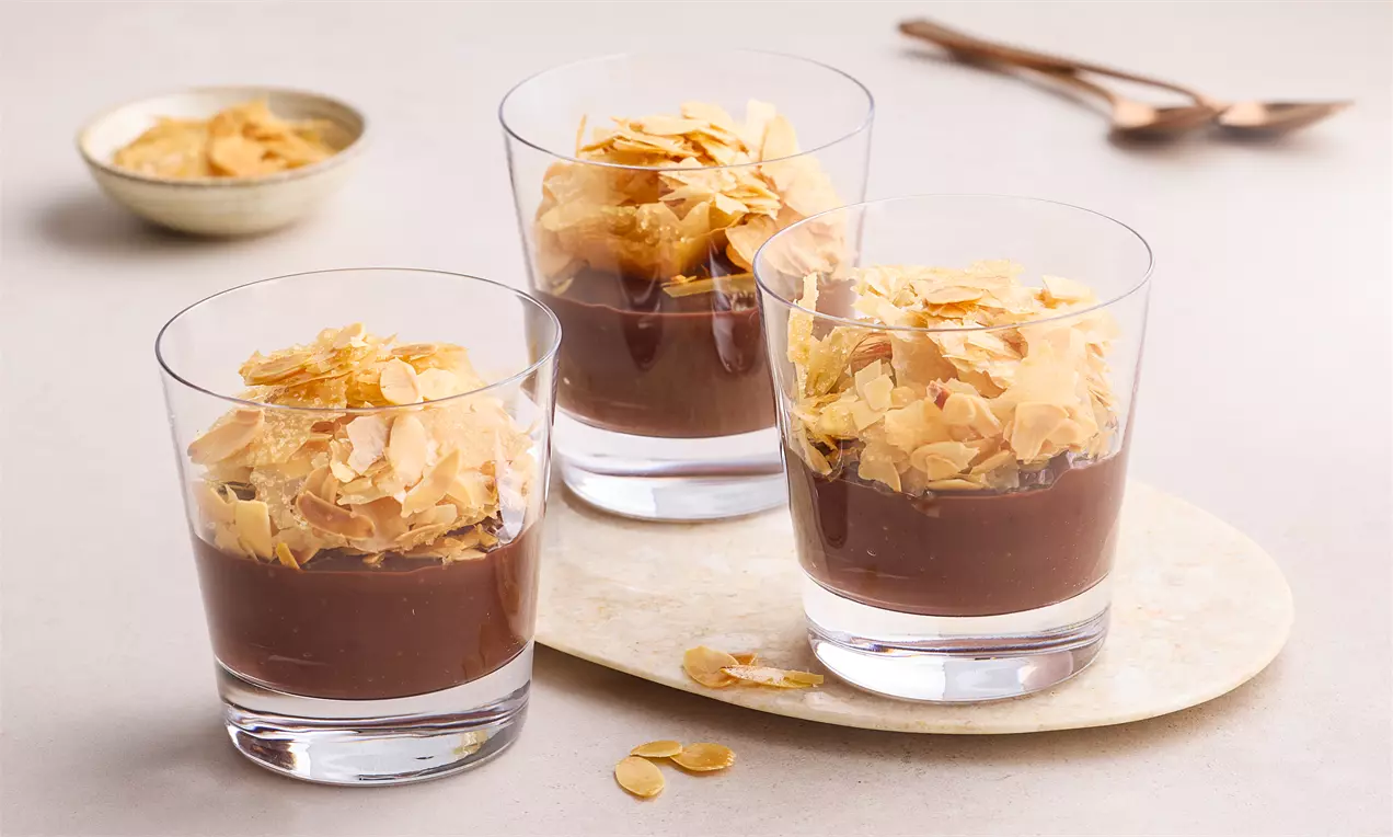 Şeffaf cam kupalarda yer alan yoğun kıvamlı çikolatalı puding üzerine serpilmiş altın rengi çıtır yufka parçaları ve file bademler