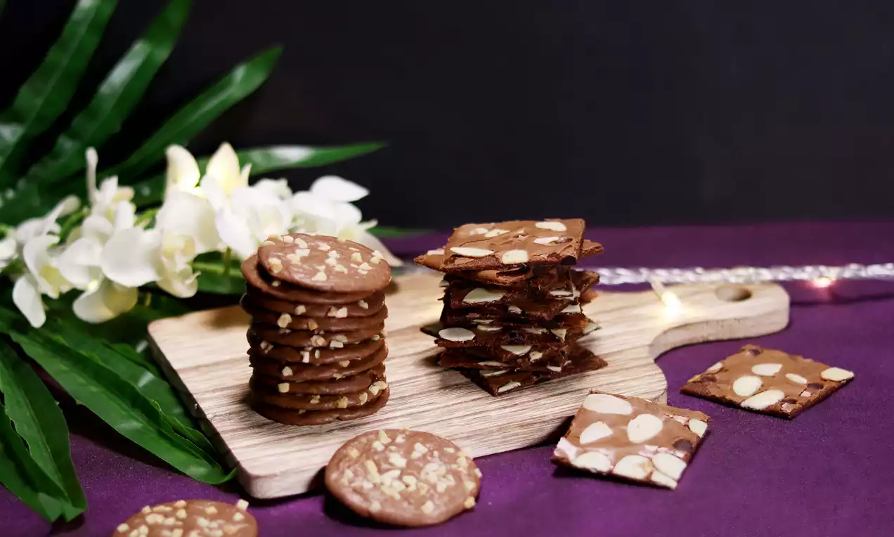 Thin round and square brownies cookies in brown tones with almond slices, stacked on a wooden board, set against a purple background with white orchids and green leaves