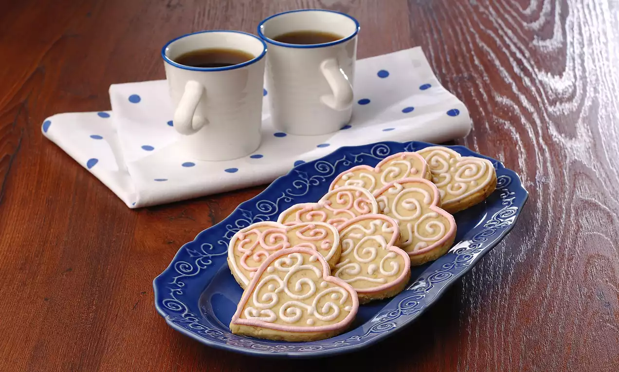 Kalp şeklinde süslenmiş kurabiyeler, mavi desenli tabakta düzenli şekilde sunulmuş, yanlarında iki fincan sıcak içecek ve beyaz puantiyeli örtü