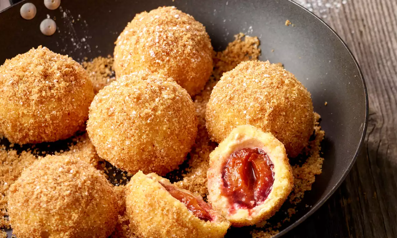 Goldbraune Zwetschkenknödel aus lockerem Topfenteig, mit buttrigen Bröseln bestreut, in einer schwarzen Pfanne angerichtet, einer aufgeschnitten