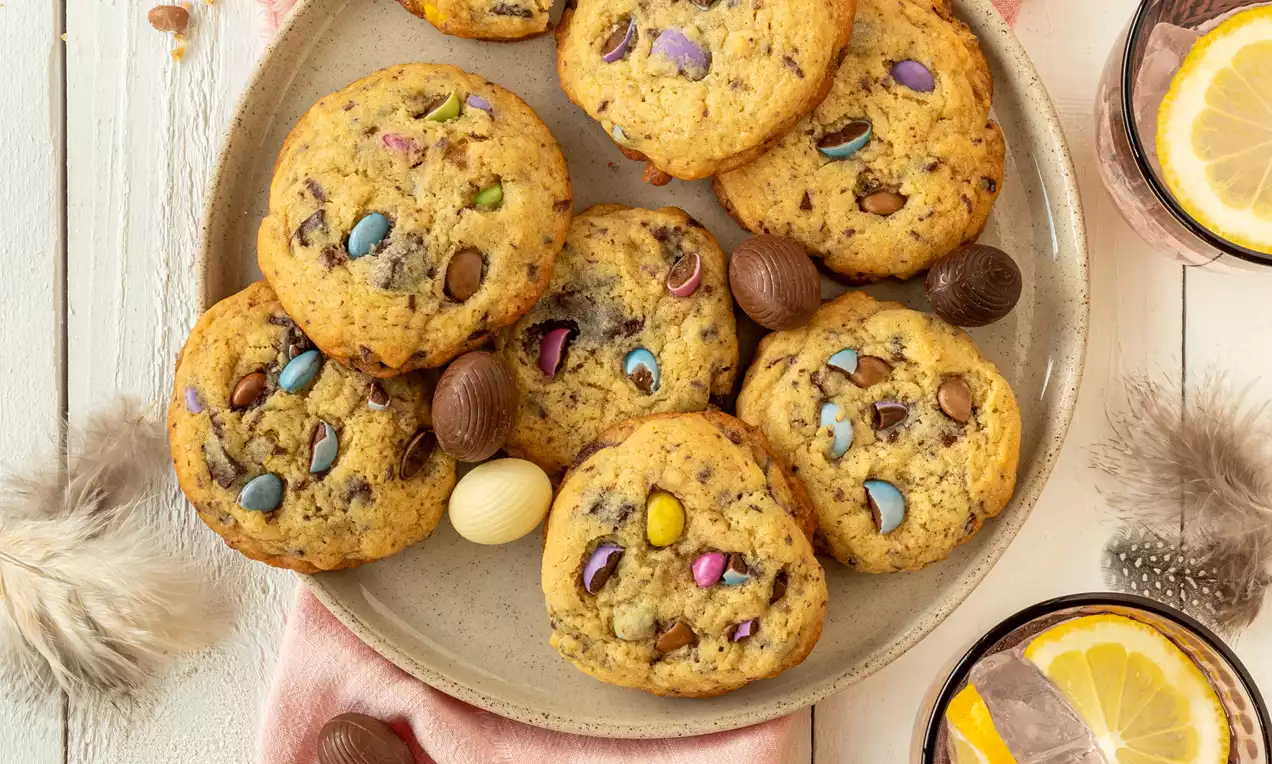 Ronde chocolate chip cookies met gekleurde chocoladepastilles op een beige bord, omgeven door paaseitjes, een roze servet en drankjes met ijs en citrus