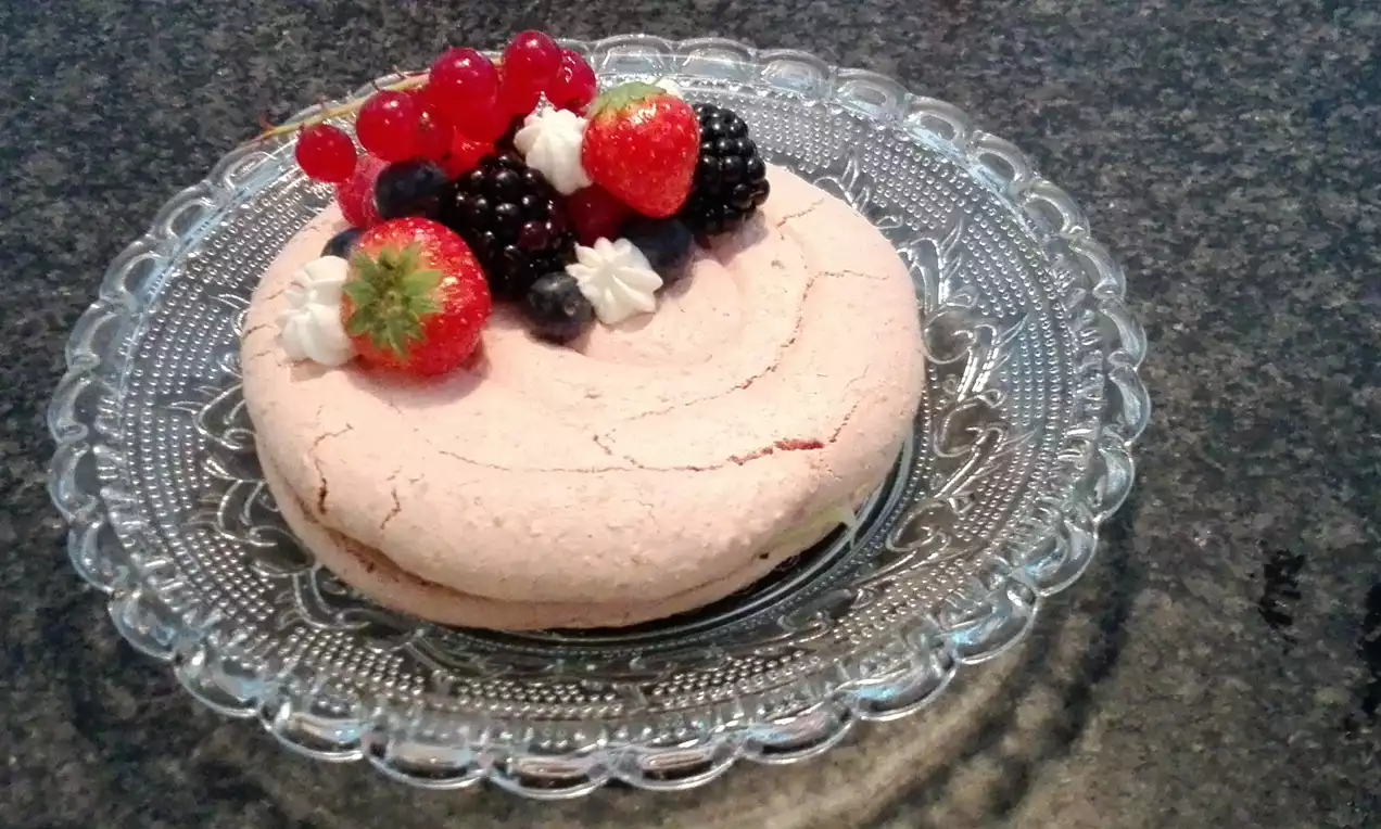 Lichtroze meringue met een korrelige textuur, versierd met rode en paarse bessen, slagroomtoefjes en aardbeien op een glazen taartbord