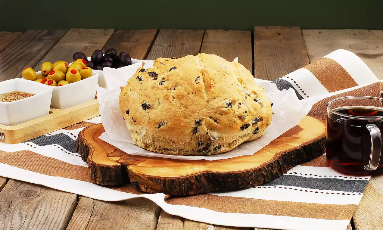 Altın rengi, kabarık ve kıtır dokulu zeytinli ekmek, rustik ahşap sunum tahtasıyla, yanındaki zeytinler ve çay eşliğinde sergileniyor