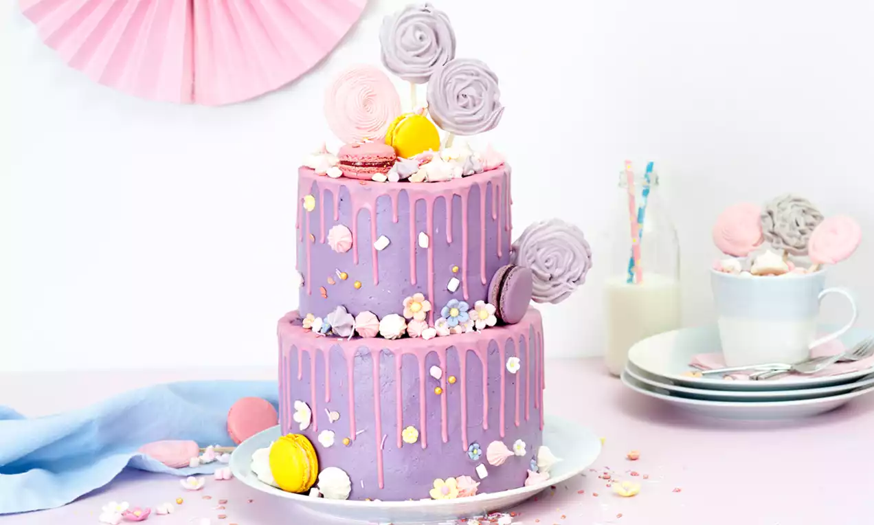 Zweistöckige, violett-rosane Torte mit Drip-Design, dekoriert mit bunten Macarons, Zuckerstangen und Zuckerblüten auf weißem Teller