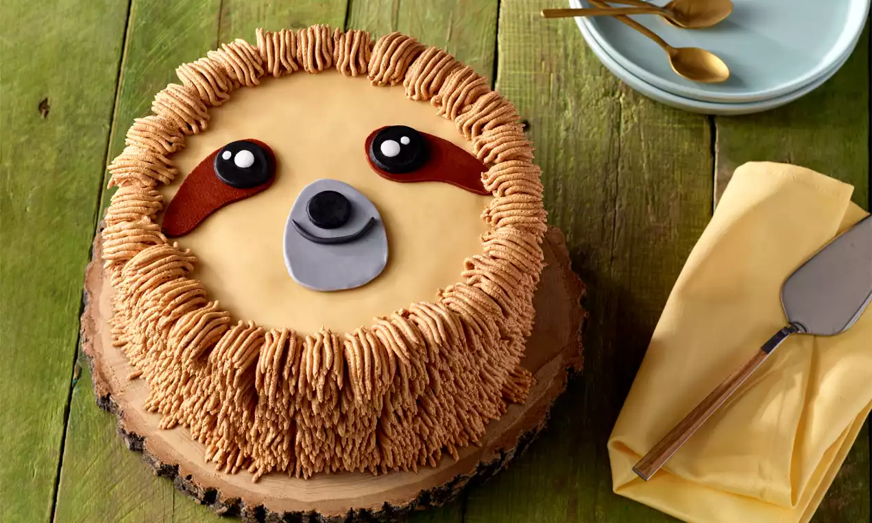 Runde Torte mit cremefarbener Fondanthülle, braunen Zuckergussstreifen als Fell und einem Faultiergesicht, auf Holzbrett angerichtet