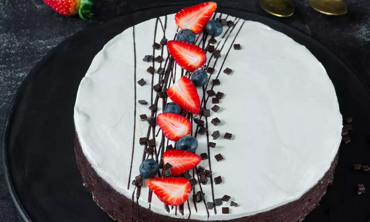 Kakaová torta s tmavým základom, hladkou bielou vrstvou, zdobená čokoládovými hoblinkami, čučoriedkami a jahodovými plátkami na čiernom tanieri