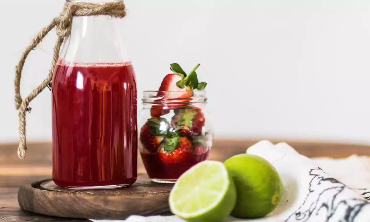 Červený sirup v lahvi se špagátovým uzávěrem na dřevěném podnosu vedle sklenice s čerstvými jahodami a dvěma limetkami na ubrusu