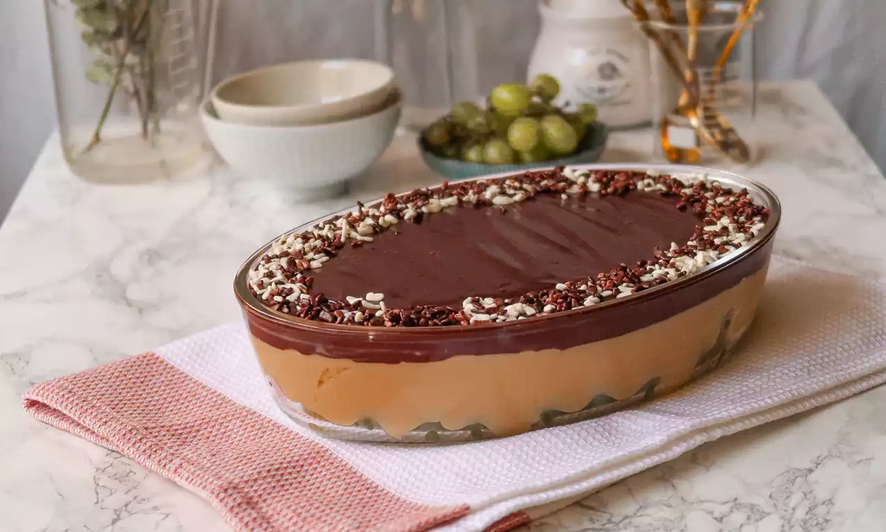 Sobremesa em camadas com creme bege, uvas verdes visíveis no fundo e cobertura de chocolate decorada com raspas brancas e escuras