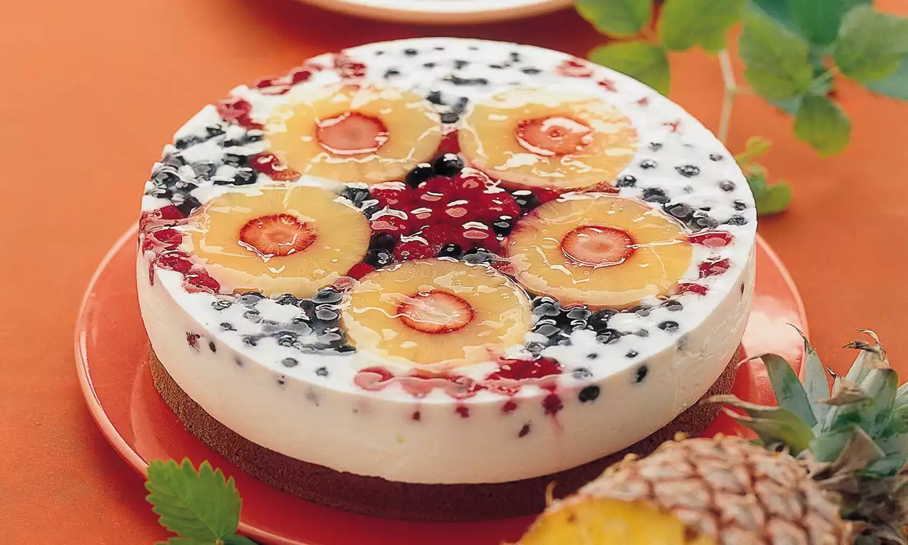Torta svijetle kreme na tamnoj podlozi, ukrašena kolutovima ananasa, jagodama, crvenim i tamnim bobicama na crvenom tanjuru