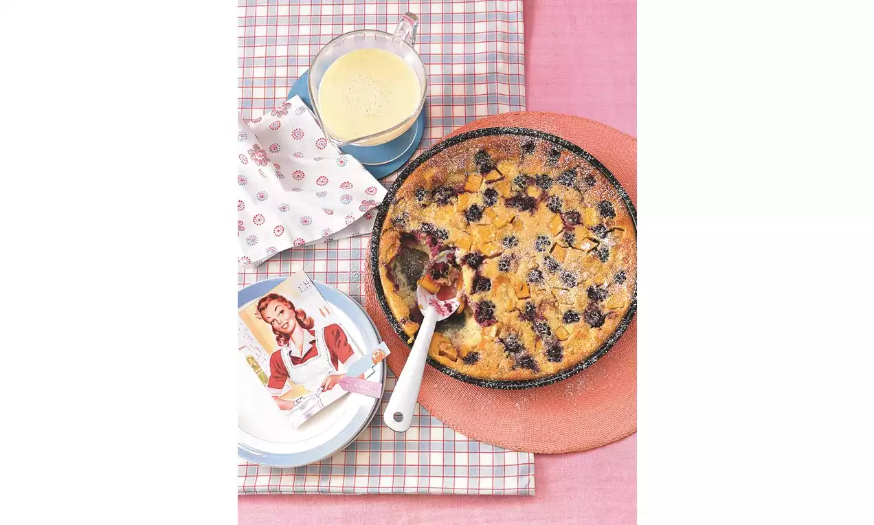 Zlatohnedý ostružinový nákyp v okrúhlej forme s jemnou práškovou cukrovou posýpkou, s naberačkou a pohárom krémovej omáčky na ružovom obruse