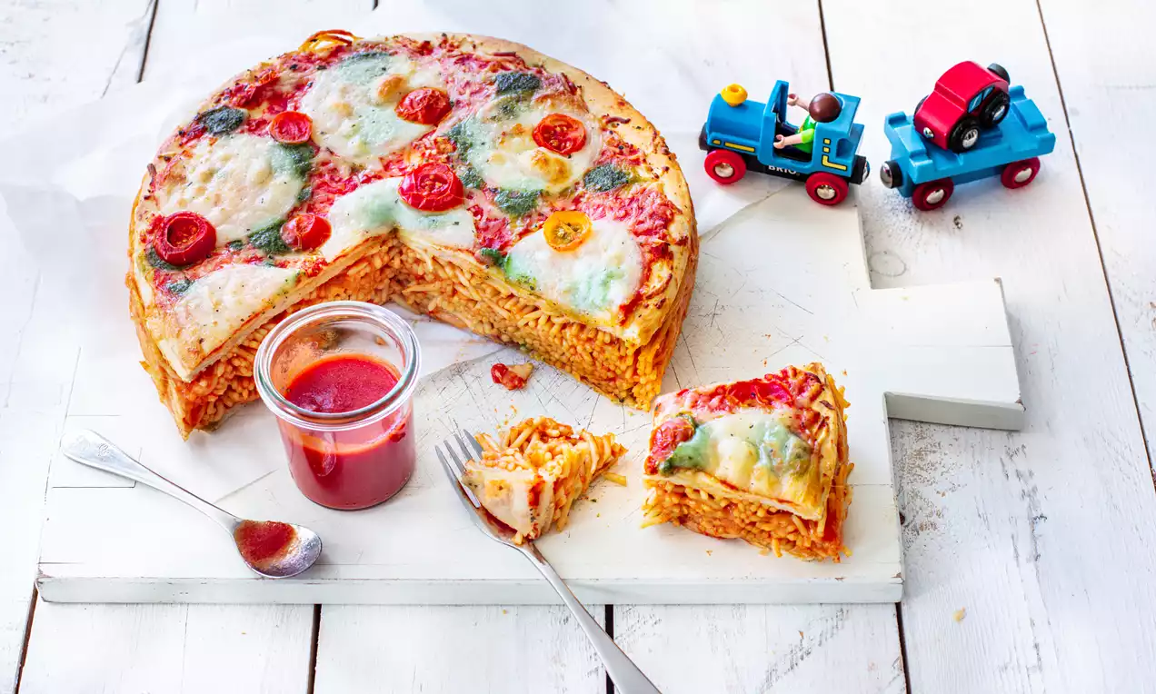 Runder Pizza-Spaghetti-Kuchen mit goldbrauner Kruste, roten Tomaten, geschmolzenem Mozzarella auf heller Holzplatte, serviert mit Tomatensoße im Glas