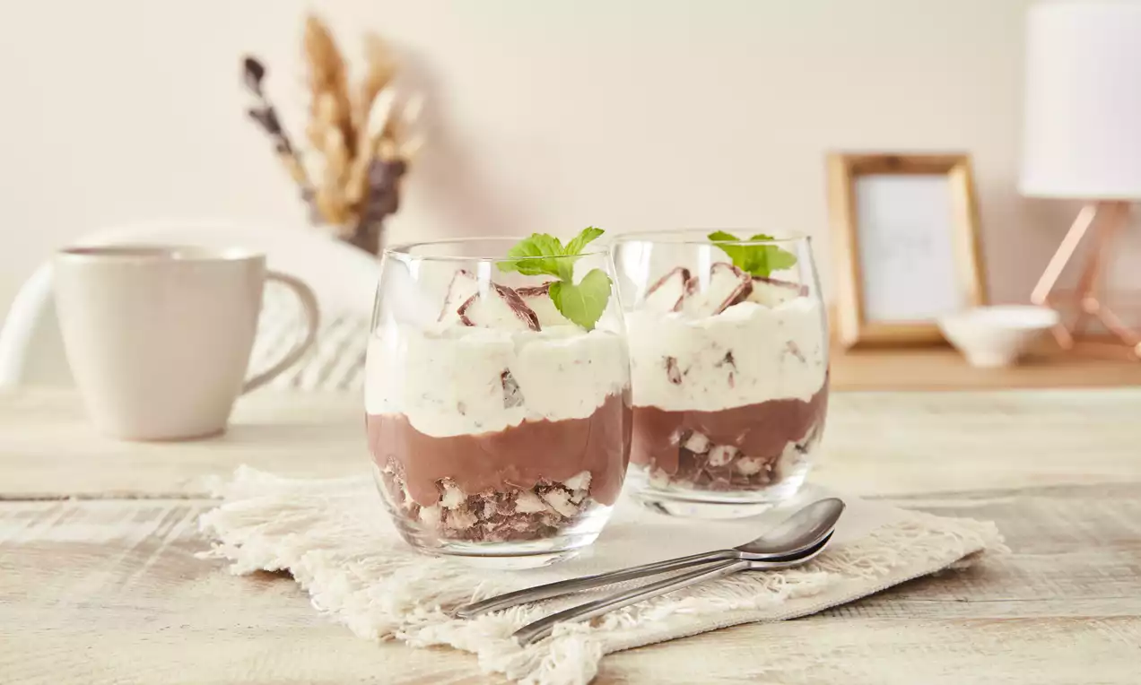 Zwei Gläser mit Schichten aus Schokocreme, knusprigen Keksstücken, weißer Kokoscreme und Bounty-Stücken, garniert mit Minzblättern