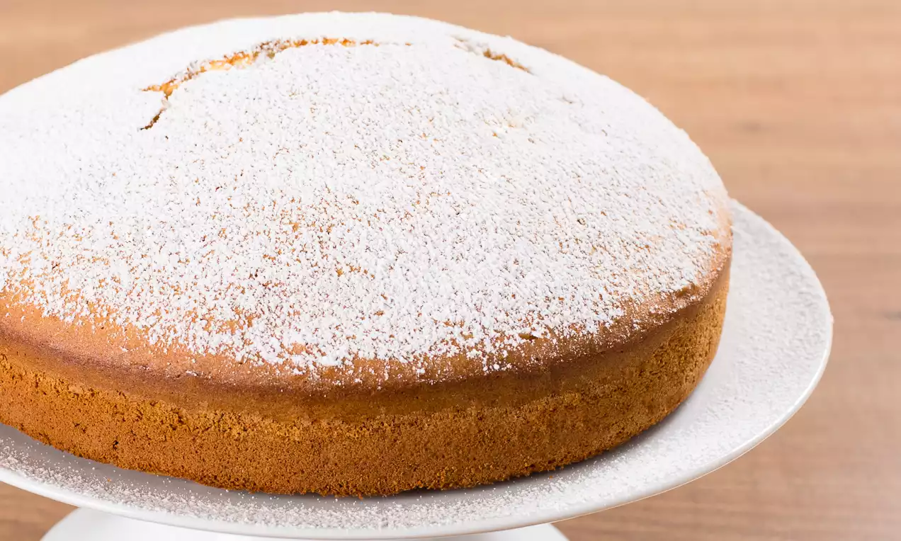 Torta alta e soffice dal colore dorato uniforme, spolverata di zucchero a velo bianco su un piatto candido e liscio