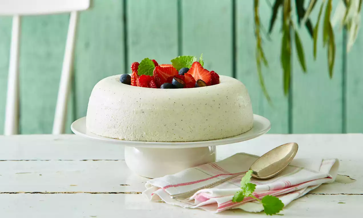 Hvid fløderand med små vaniljeprikker på et hvidt kagefad, toppet med røde bær, blå bær og grønne mynteblade