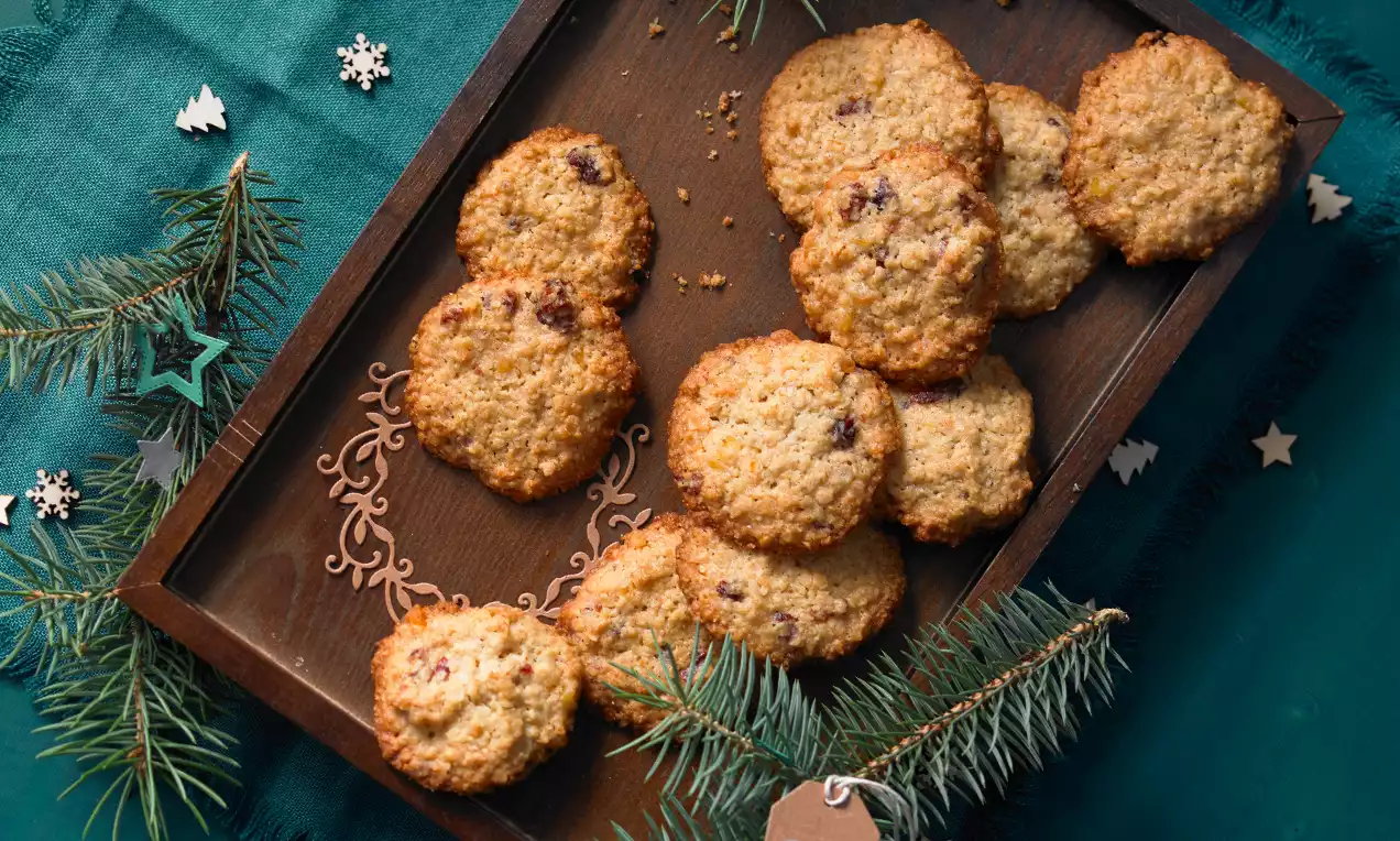 Goldbraune Haferflockenplätzchen mit leichter Krümeltextur und vereinzelt dunklen Trockenfrüchten auf einem Holztablett mit Tannenzweigen drapiert