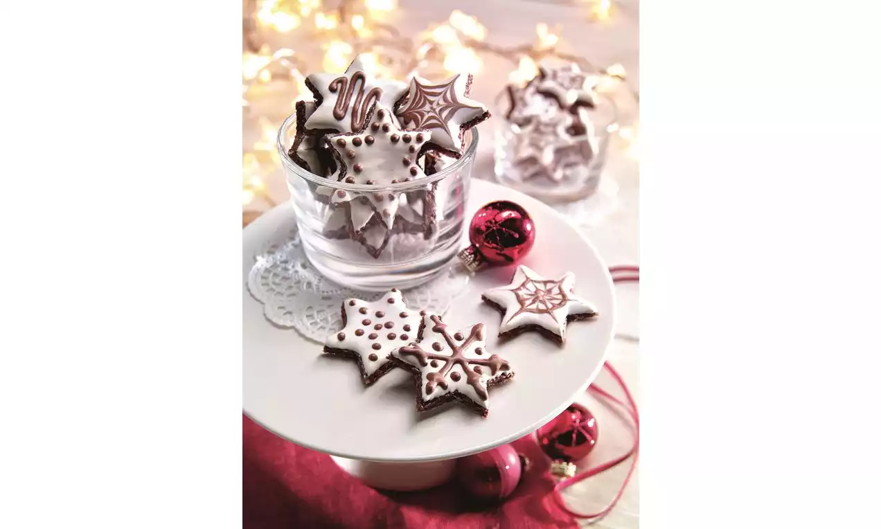 Mandľové kakaové hviezdičky s bielou lesklou polevou zdobené čokoládovým vzorom, rozložené na sladkom tanieri vedľa červených vianočných ozdôb