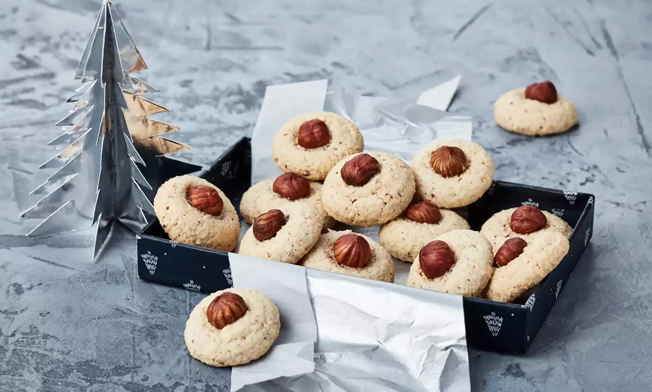 Biscotti rotondi dal colore chiaro con consistenza leggermente granulosa, decorati con nocciole intere e disposti in una scatola su carta argentata