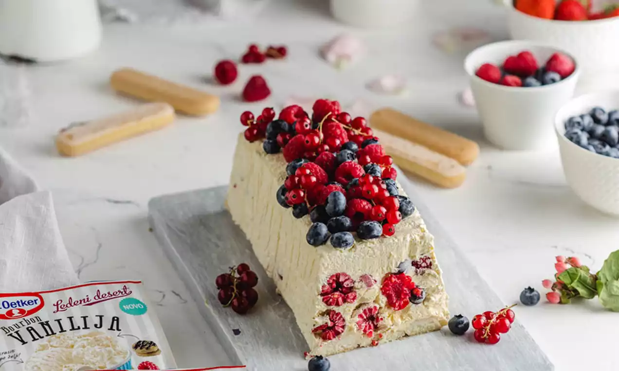 Semifreddo svijetložute boje pravokutnog oblika, posut svježim malinama i borovnicama na sivom pladnju okružen voćem i dodatcima