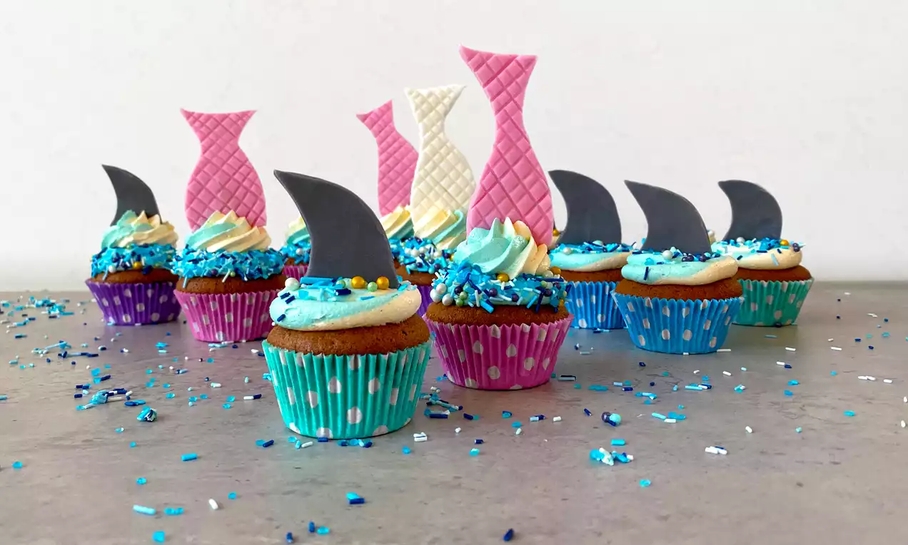 Cupcakes med farverigt papirmuffinsform, blå krymmel, frosting og pynt af grå hajfinner og lyserøde eller hvide havfruehaler