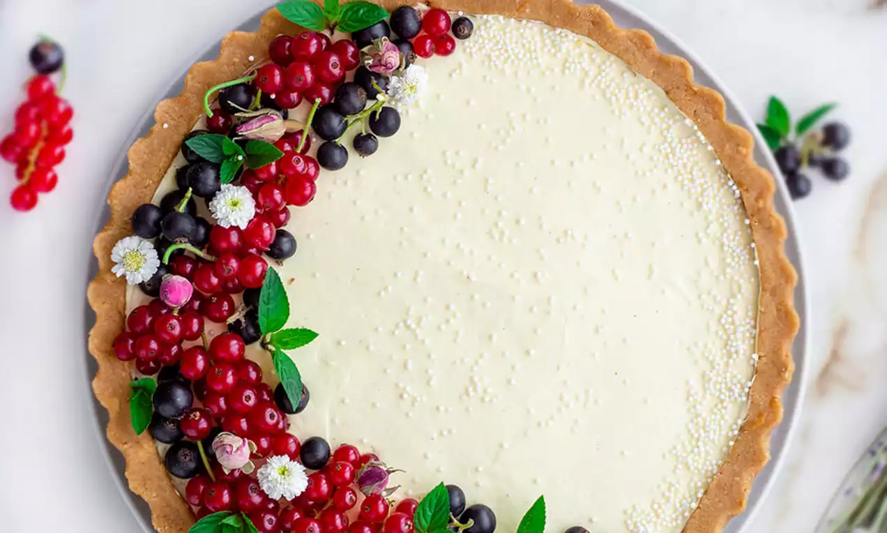 Tart s glatkom svijetložutom vanilija kremom, okružen hrskavim smeđim rubom, ukrašen bobičastim voćem, cvjetovima i svježim listićima na bijelom tanjuru