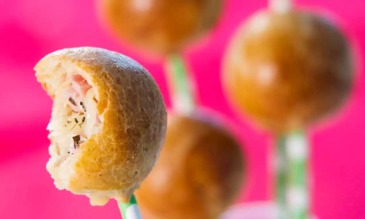 Bolinhas douradas sobre palitos verdes com recheio aparente de textura cremosa e detalhes de ervas ao fundo rosa vibrante