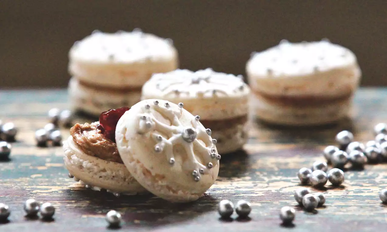 Smetanově bílé makronky s hladkou texturou zdobené stříbrnými perličkami a vločkovým vzorem, uspořádané na dřevěném povrchu mezi stříbrnými dekoracemi